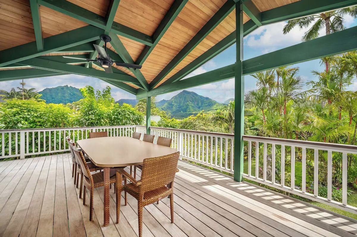 The covered upper deck at Mala Puʻa Ma Ke Kai, set with a long dining table for ten, looking out over palms to the green Hanalei mountains.